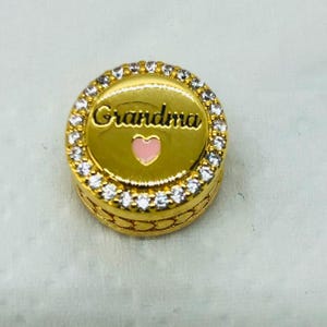 May include: A gold-colored charm with the word "Grandma" in cursive script and a pink heart. The charm is round, with a border of clear, sparkling stones. The charm is likely for a bracelet or necklace.