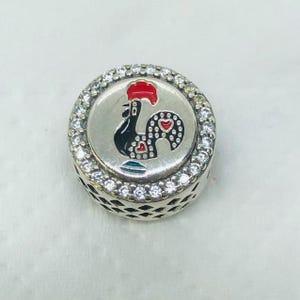 May include: A round, silver charm with a detailed design. The charm features a black rooster with a red comb and heart-shaped details. The charm is surrounded by a ring of clear, sparkling stones, adding a touch of elegance to the piece.