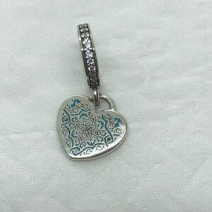 May include: Silver heart-shaped charm with an intricate blue and silver floral design. The charm has a decorative bail with clear stones. The heart pendant is suspended from a silver loop.