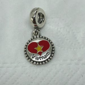 May include: A silver charm featuring a red heart and yellow star design, with the word "Vietnam" in silver lettering. The charm has a beaded border and a silver loop for attaching to a bracelet or necklace.