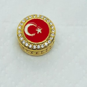 Puede incluir: Un pin redondo dorado con un centro rojo que presenta un diseño de luna creciente y estrella blancas, rodeado por un anillo de piedras preciosas transparentes. El pin tiene un borde texturizado.