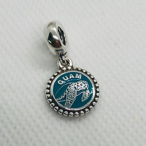 May include: A silver-toned metal charm featuring a circular pendant with a beaded edge. The centre is filled with teal enamel, displaying a white sea turtle graphic and the word GUAM arched above the turtle in white capital letters.