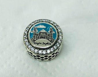 Puede incluir: Charm de plata con una ilustración detallada de la mezquita de Santa Sofía sobre un fondo de cielo azul. El charm está rodeado por una fila de piedras claras y brillantes. El texto "Hagia Sophia" es visible.