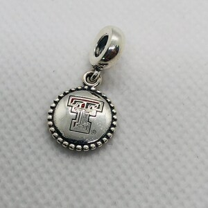 May include: A silver-toned metal charm featuring a circular pendant with a beaded edge. The center of the pendant displays a stylized letter T logo with red accents and a small registered trademark symbol. The charm includes a cylindrical bail.