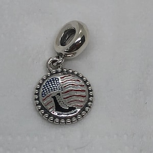 Pode incluir: Um berloque de prata com um pingente redondo com um design de bandeira americana vermelha, branca e azul e uma bota militar preta. O berloque tem uma borda com contas e uma argola para prender a uma pulseira ou colar.