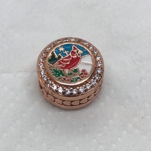 May include: A rose gold charm with a detailed design. The charm features a red cardinal bird on a branch, surrounded by a ring of clear crystals. The background includes a winter scene with a blue sky and green grass.