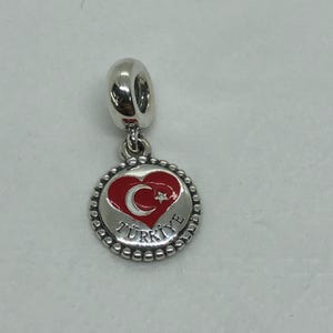 Puede incluir: Un charm de plata con un corazón rojo con una luna creciente y una estrella blancas. El charm está rodeado por un borde con cuentas y la palabra "TURKIYE" está inscrita alrededor del borde.