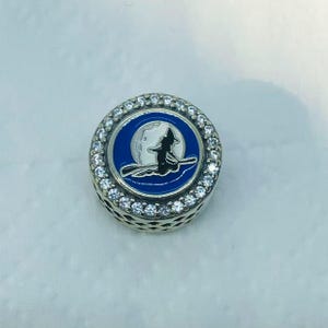 May include: A silver charm with a blue and white enamel design featuring a witch flying in front of a moon. The charm is round, with a ring of clear stones around the edge.