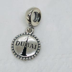 May include: Silver charm with a round pendant featuring the word "DUBAI" and a black silhouette of a tall building. The pendant is surrounded by a beaded border and attached to a silver loop.