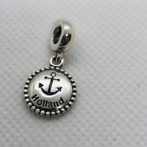 May include: A silver-toned metal charm featuring a circular pendant with a beaded edge. The center displays a black engraved anchor symbol above the word Holland in black lettering. The charm includes a cylindrical bail for attachment to a bracelet.