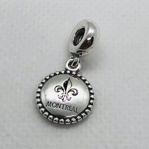 Pode incluir: Um berloque de metal prateado com um pingente circular e borda de contas. O centro exibe um símbolo de flor-de-lis preto acima do texto MONTREAL. O berloque inclui uma argola polida no topo para fixação em uma pulseira ou colar.