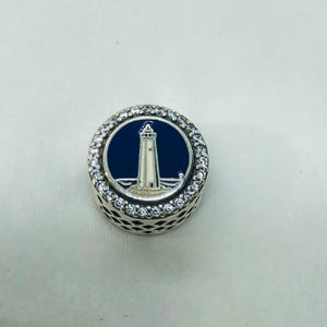 May include: A round silver charm with a lighthouse design. The charm features a white lighthouse on a blue background, surrounded by clear crystals. The charm has a detailed, openwork design on the sides.