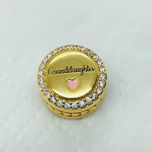 May include: A gold-colored charm with the word "Granddaughter" in black script and a pink heart. The charm is round, with a ring of clear crystals around the edge.