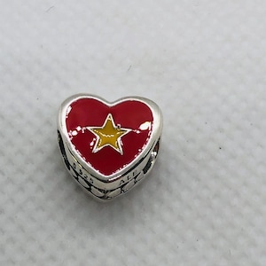 Könnte beinhalten: Ein herzförmiger silberner Charm mit einer leuchtend roten Emaille-Einlage und einem gelben Stern in der Mitte. Die Seite des Metallanhängers ist mit S925 und ALE graviert, und der äußere Rand zeigt ein wiederkehrendes Kreismuster in poliertem Silber.