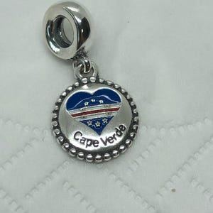 May include: A silver charm with a blue and white heart-shaped design, featuring the Cape Verde flag. The charm has a beaded border and the words "Cape Verde" are inscribed on the bottom.