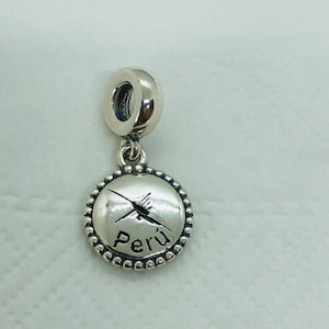 May include: A silver charm with a round pendant featuring a stylized hummingbird design and the word "Perú". The charm has a beaded border and a loop for attaching to a bracelet or necklace.