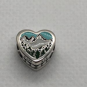 May include: A heart-shaped silver charm featuring an enamel inlay of a mountain range with white snow-capped peaks, dark green pine trees, and a light blue sky. The side of the charm is engraved with the text 925 and ALE in black lettering.