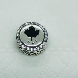 May include: A round silver charm with a black maple leaf design and the word "Banff" in script. The charm is encircled by a row of clear, sparkling stones, adding a touch of elegance to the piece.