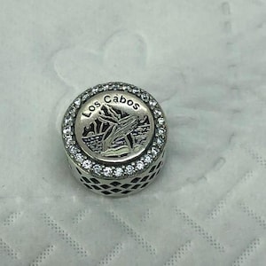 May include: A silver-toned charm with the words "Los Cabos" encircling a whale design. The charm is round, with a ring of clear stones around the top. The sides of the charm have a decorative pattern.