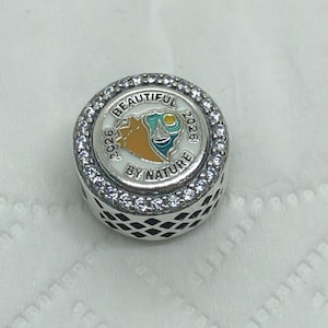 May include: A round silver charm with a white enamel center featuring a colorful landscape design and the words "BEAUTIFUL BY NATURE 2026". The charm is encircled by clear crystals.