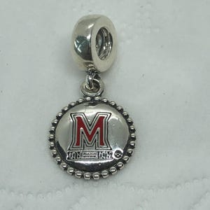May include: A silver-toned metal charm featuring a circular pendant with a beaded edge. The center displays a red and white letter M logo with small text underneath. The charm includes a cylindrical bail at the top for attachment to a bracelet.