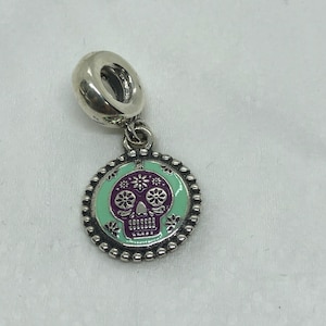 May include: A silver charm with a Day of the Dead sugar skull design. The charm features a round pendant with a mint green background, a purple skull, and floral accents. The pendant is surrounded by a beaded border.
