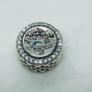 May include: A round, silver charm with a detailed landscape scene in blue and black enamel. The charm is encircled by a row of clear, sparkling crystals. The sides of the charm have a decorative, openwork design.
