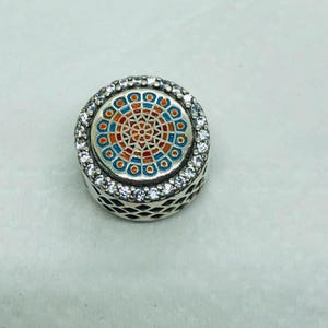 Könnte beinhalten: Ein runder Silberanhänger mit einem farbenfrohen Buntglasdesign in Rot-, Orange- und Blautönen. Der Anhänger ist von einer Reihe klarer, funkelnder Steine umgeben. Die Seiten des Anhängers haben ein dekoratives, durchbrochenes Muster.