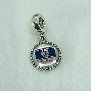 May include: A silver charm with a round pendant featuring the Belize flag. The flag design includes a blue background with a red stripe and the Belize coat of arms. The word "Belize" is written in white. The charm has a beaded border and a loop for attachment.
