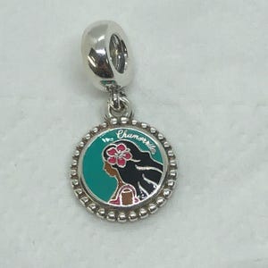 May include: A silver charm with a round pendant featuring a woman's profile, a flower, and the word "Chamorrrita". The pendant has a teal background and a beaded border. The charm has a loop for attaching to a bracelet or necklace.