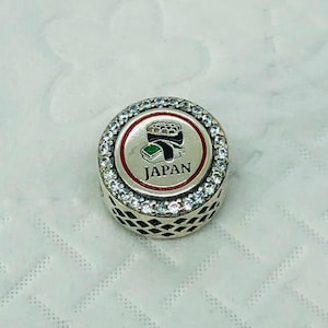 May include: Silver-toned charm with a sushi design. The charm features a central circular design with a sushi illustration and the word "JAPAN" in black text. The charm is surrounded by clear, sparkling stones.
