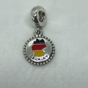 May include: A silver charm with a loop at the top and a round pendant. The pendant features a map of Germany in black, red, and yellow, with the word "GERMANY" in a circular border. The charm is likely for a bracelet or necklace.