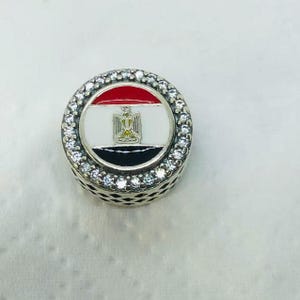 May include: A round charm with the flag of Egypt. The charm features a red, white, and black design with the Egyptian coat of arms. The charm is surrounded by a ring of small, clear crystals and has a silver-colored base.