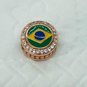 May include: A round, rose gold-colored charm with a Brazilian flag design in the center. The flag features a green background, a yellow diamond, and a blue circle with white stars. The charm is encircled by clear crystals.