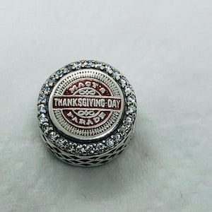 May include: A round, silver charm with the text "Macy's Thanksgiving Day Parade" in red and white. The charm is encircled by a row of clear, sparkling stones. The design is detailed and festive, perfect for a holiday-themed bracelet or necklace.