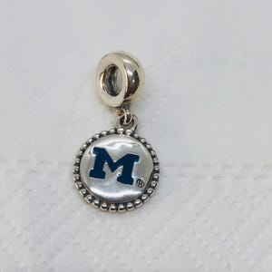 Puede incluir: Un charm de plata con un colgante redondo con una "M" azul marino sobre fondo blanco. El colgante está rodeado por un borde de cuentas y cuelga de un lazo plateado. El charm está diseñado para una pulsera o un collar.