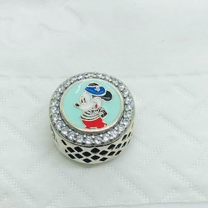 May include: A round silver charm bead featuring a central light blue enamel inlay with a cartoon mouse character wearing a blue hat, a striped shirt, and red trousers. The outer edge is encrusted with clear stones and a black lattice pattern.