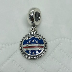 May include: A silver charm with a round pendant featuring the Cape Verde flag design. The pendant has a blue background with white stars and a red and white stripe. The words "Cape Verde" are written around the edge.