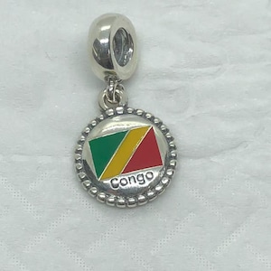 Puede incluir: Un charm de plata con un colgante redondo que presenta la bandera del Congo. El diseño de la bandera incluye triángulos verdes, amarillos y rojos, con la palabra "Congo" en blanco. El charm tiene un borde con cuentas y un lazo.