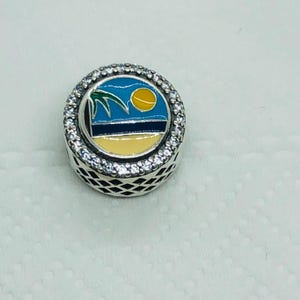 May include: A round silver charm with a beach scene design. The charm features a blue sky, yellow sun, palm tree, and a sandy beach. The charm is surrounded by a ring of small, clear stones.