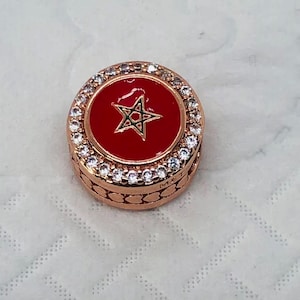 May include: Rose gold-toned charm with a red enamel center featuring a green star design. The charm is encircled by a row of clear, sparkling crystals. The charm has a detailed, textured band.