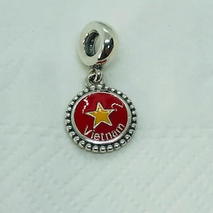 May include: A silver charm with a red and yellow enamel design. The charm features a red circle with a yellow star and the word "Vietnam" in white. The charm is round with a beaded edge and a silver loop.