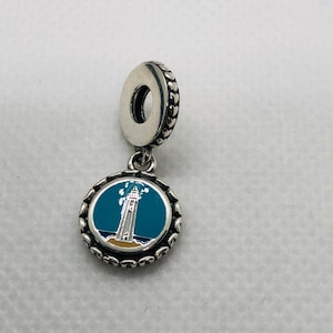 May include: A silver dangle charm featuring a circular pendant with a teal enamel inlay depicting a white lighthouse on a tan shore against a dark blue sea. The charm has a textured border of small hearts and a silver bail with a heart pattern.