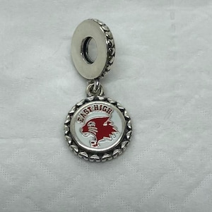 Puede incluir: Un charm de plata con un colgante redondo con las palabras "East High" y un logotipo de puma rojo. El charm tiene un borde decorativo con cuentas y un lazo para sujetarlo a una pulsera o collar.