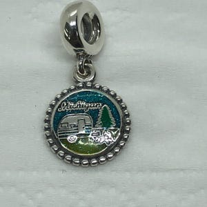 Puede incluir: Un charm de plata con un colgante redondo con una caravana, un árbol y la palabra "Michigan". El colgante tiene un fondo azul y verde y un borde con cuentas. El charm tiene un lazo en la parte superior.
