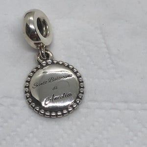 May include: A silver charm with a round pendant. The pendant has a beaded edge and engraved text that reads "Santo Battesimo di Amelia". The charm has a loop at the top for attaching to a bracelet or necklace.