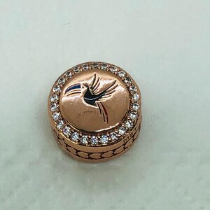 May include: A circular, rose gold charm featuring a bird design. The bird is black with red and white details, and a blue and white flag. The charm is encircled by a ring of small, clear crystals.