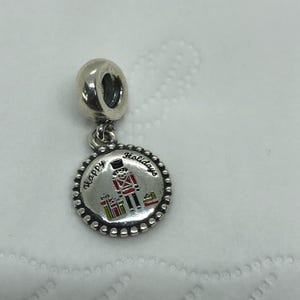 May include: A silver-toned metal charm featuring a circular pendant with a beaded edge. The center displays a colorful nutcracker figure and wrapped gift boxes, with the engraved text "Happy Holidays" arched across the top of the design.