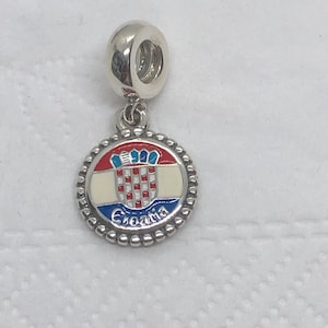 May include: A silver charm with a round pendant displaying the Croatian coat of arms. The pendant features a red and white chequered shield, a blue and white crown, and the word "Croatia" in blue. A souvenir or jewellery item.