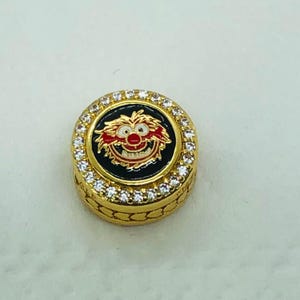 Puede incluir: Un pin circular de color dorado con un diseño de personaje de dibujos animados. El pin presenta un centro negro con una cara de personaje roja y blanca. El anillo exterior está incrustado con pequeños cristales transparentes.